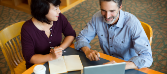 Two library professionals sitting at a table with a laptop, book, and coffee, talking. 